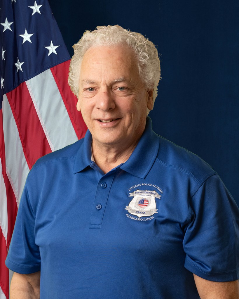 A portrait of Michael Szaz in front of an American Flag and blue background.